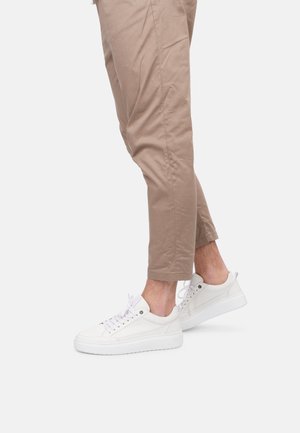Weiße Ledersneaker mit runder Zehenpartie, flacher weißer Sohle, metallischen Ösen und weißen Schnürsenkeln, getragen mit hellbeigen, schmal geschnittenen Hosen.