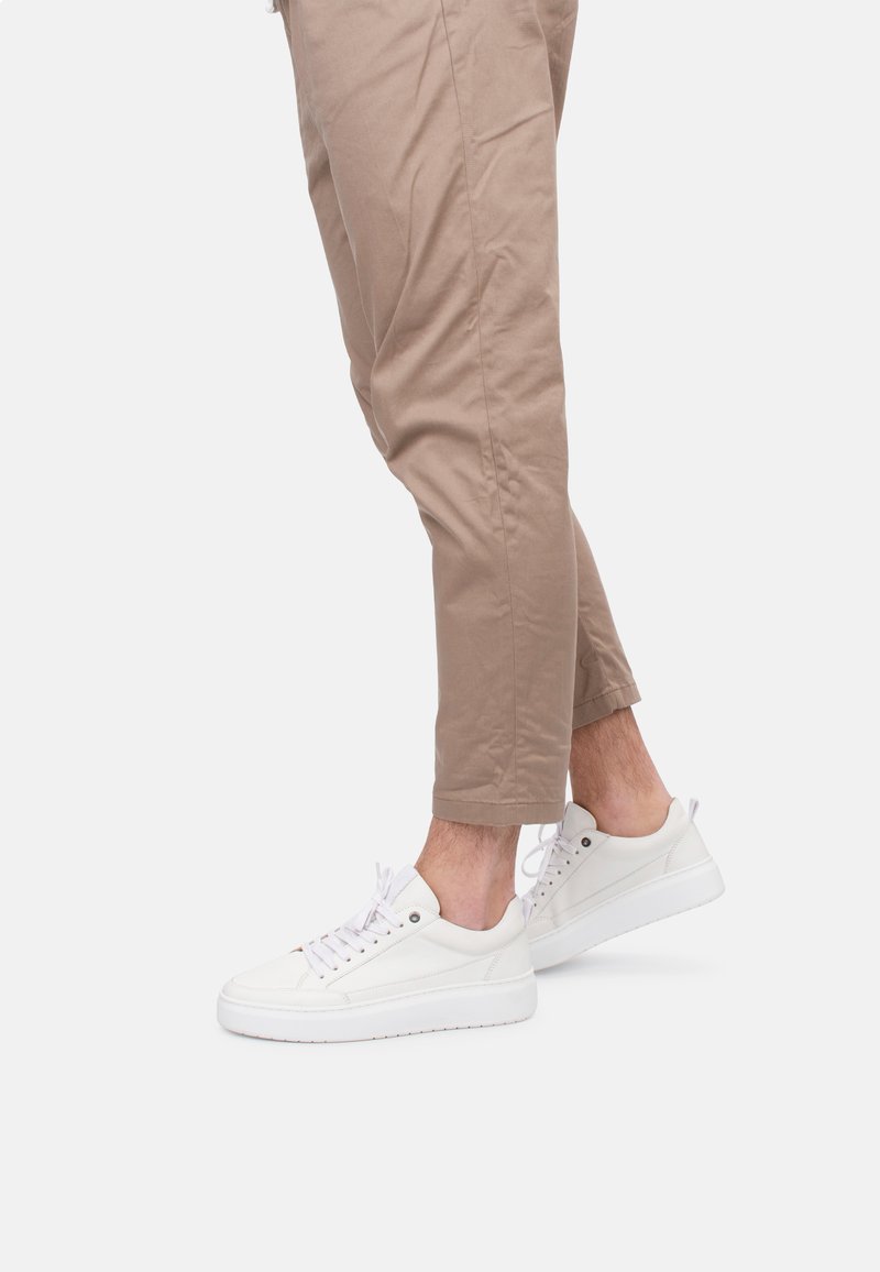 Sneakers bianchi in pelle con punta rotonda, suola piatta bianca, occhielli metallici e lacci bianchi, indossati con pantaloni tapered beige chiaro.