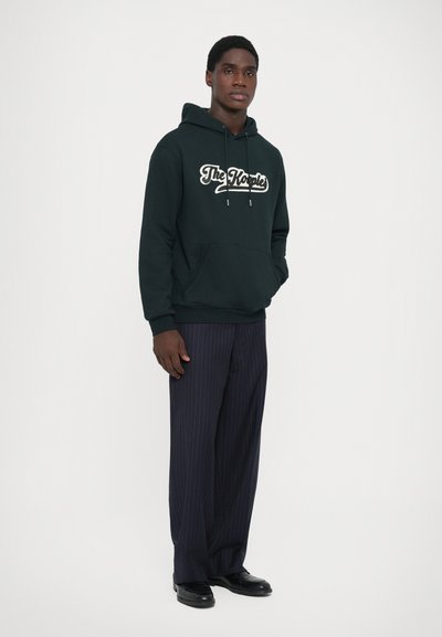 Dark green hoodie with a white script logo, kangaroo pocket, and drawstring hood, paired with navy pinstriped trousers and black shoes.