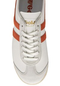 Sneaker bianca con strisce in suede arancione, lacci in tessuto e logo dorato. La punta arrotondata e la suola testurizzata offrono elementi di design sportivi.