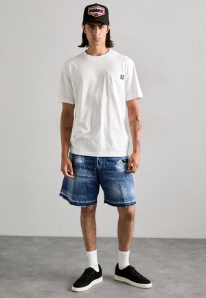 T-shirt bianco a maniche corte con taschino sul petto, abbinato a pantaloni shorts in denim strappati. Un cappellino nero, scarpe da ginnastica nere e calze bianche completano l'outfit.