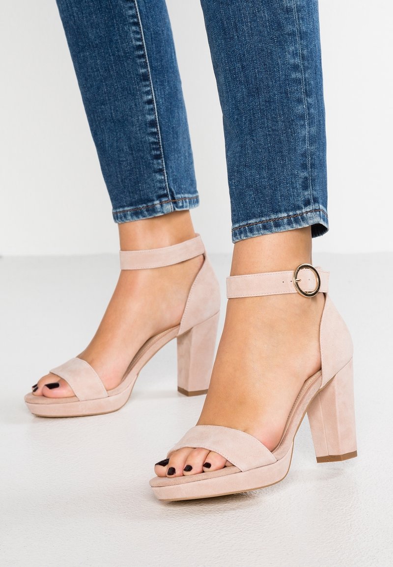 Žena v světle růžových semišových sandálech na vysokém podpatku s kotníkovými pásky a tmavým lakem na nehty, v modrých džínách.