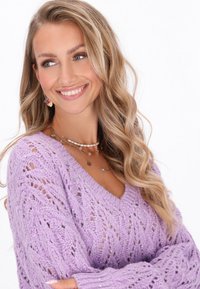 Pull en laine lavande avec un col en V, présentant un design en dentelle et des accents argentés. Des bijoux en or et perles superposés ornent le cou.