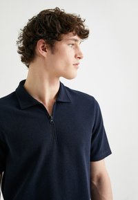 Marinblå piké med kort ärm i texturerad tyg med dragkedja i kragen. Enkel design, slim fit och utan synliga logotyper eller mönster.