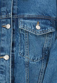 Denim jacket in light blue, featuring silver snap buttons, a chest pocket with a flap, and brown stitching along the seams.