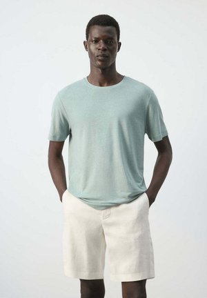Joven de piel oscura con una camisa de manga corta verde claro y pantalones cortos beige, de pie con las manos en los bolsillos frente a un fondo liso.