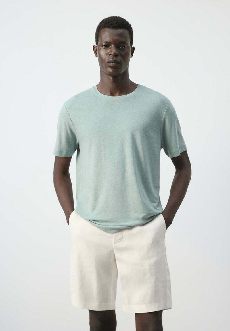 Jeune homme à la peau foncée portant une chemise à manches courtes vert clair et un short beige, debout les mains dans les poches devant un fond uni.