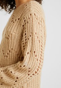 Maglione beige lavorato a maglia con un design a punto aperto, maniche lunghe e un motivo testurizzato con fili sciolti, in un taglio rilassato.