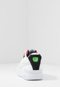 Baskets blanches Puma avec un patch noir au talon, logo vert, doublure intérieure rouge et semelle blanche texturée, vues de dos sur une surface blanche.