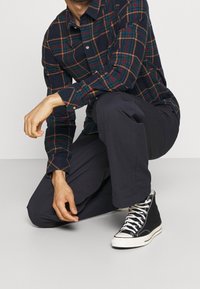 Camisa de franela de base azul marino, con un patrón de cuadros en verde y naranja. Combinada con pantalones oscuros y zapatillas altas negras con detalles en blanco.
