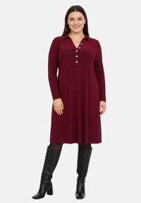 Robe bourgogne à manches longues avec col et patte de boutonnage, en tissu doux, associée à des bottes noires jusqu'aux genoux.