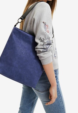 Femme portant un sweat-shirt gris clair avec une broderie bleue et un jean, portant un grand sac à bandoulière bleu texturé avec une sangle noire.