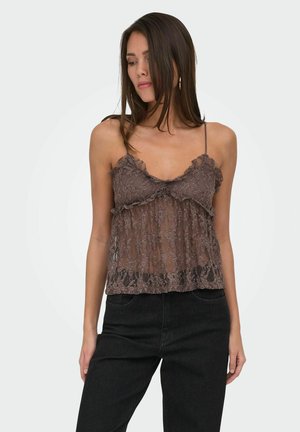 Kvinde iført brun blondeærmeløs top og sorte bukser, stående mod en ensartet lys baggrund, kigger ned og til siden.