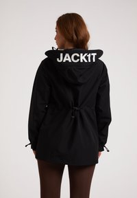 Svart vattentät jacka med dragsko i midjan, justerbar huva och stor "JACK1T"-logotyp på baksidan. Har en slät, matt yta.