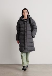 Abrigo de plumas en gris oscuro con capucha, con cierre de botones, mangas largas y detalles acanalados. Llevado sobre pantalones verde claro.