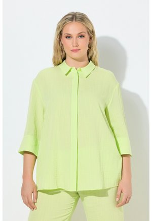Vrouw met blond haar die een loszittend, limoen groen textuur patroon overhemd met knopen en bijpassende broek draagt, staand tegen een witte achtergrond.