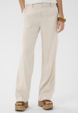 Naine kannab beeži laia säärega pükse, valget toppi, puidust platvormsandaale ja merevaiguvärvi käevõru, seisab valgel taustal käed taskus.