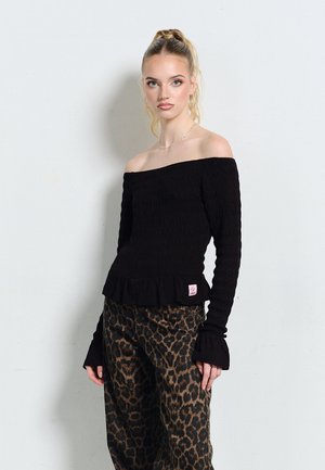 Młoda kobieta o blond kucyku, ubrana w czarny, odkrywający ramiona top z długim rękawem oraz spodnie w panterkę, stoi na tle jasnoszarej ściany.