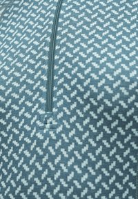 Tissu tricot bleu et blanc avec une fermeture éclair diagonale, montrant un motif géométrique répétitif et un petit carré cousu sous la fermeture éclair.
