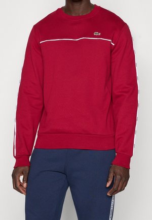 Mężczyzna w czerwonej bluzie z okrągłym dekoltem, ozdobionej białym przeszyciem i logo na piersi, w zestawie z granatowymi spodniami z bocznymi paskami.