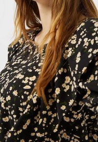 Blouse noire à motif floral avec un petit motif de marguerites blanches et jaunes. Le tissu est léger avec une texture douce et une coupe décontractée.