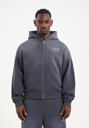 Modello maschile con pelle scura e capelli intrecciati indossa una felpa con cappuccio grigio scuro con zip e pantaloni della tuta abbinati con logo AVX sul petto e sulla coscia.