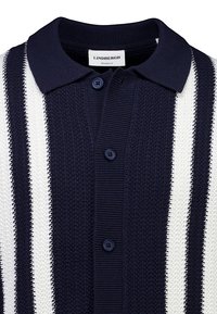 Polo en maille bleu marine avec rayures blanches verticales, boutonné sur le devant, et étiquette "LINDBERGH" sur le col, style coupe décontractée.