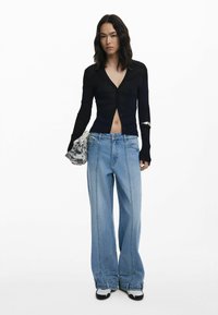 Zwarte geribbelde cardigan met V-hals en knopen, gecombineerd met lichtblauwe wijde jeans. Model houdt een bedrukte tas vast en draagt zwart-witte schoenen.
