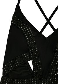 Prenda negra con tirantes entrecruzados, adornada con un tejido negro texturizado y tachuelas plateadas para un detalle adicional. Fondo de tela suave.