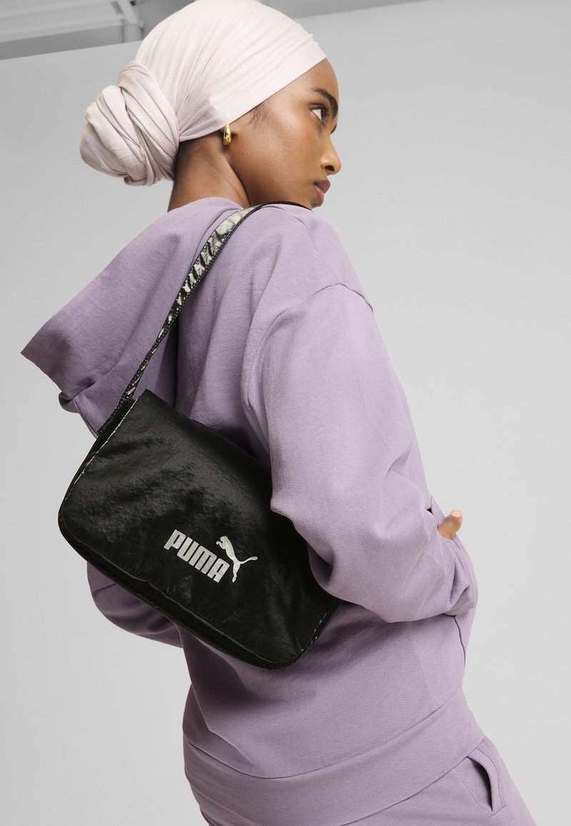 Borsa a spalla nera con texture lucida, caratterizzata dal logo bianco "PUMA" e da una silhouette animale. Indossata con una felpa leggera viola e una tracolla decorata.