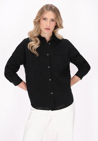 Cămașă neagră cu nasturi, cu mâneci lungi, av�ând o textură subtilă, un buzunar pe piept și nasturi din argint. Este asortată cu pantaloni albi.