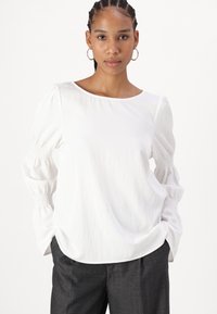 Blusa bianca con scollo rotondo e maniche a sbuffo, caratterizzata da dettagli smock ai polsini. Abbinata a pantaloni grigi scuri.