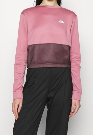 Kobieta nosząca różowo-czarny crop top North Face z długim rękawem w połączeniu z czarnymi spodniami z wysokim stanem, stojąca na jednolitym tle.