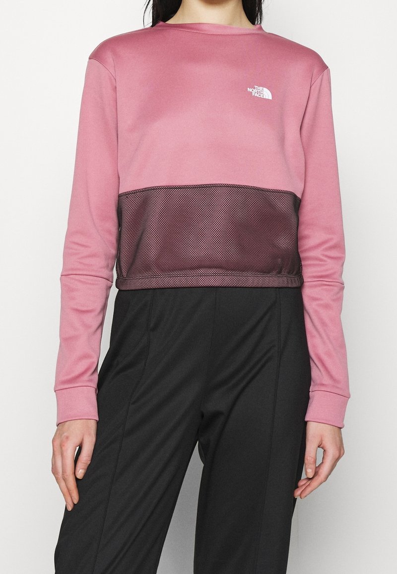 Femme portant un crop top North Face rose et noir à manches longues, associé à un pantalon noir taille haute, debout devant un fond uni.
