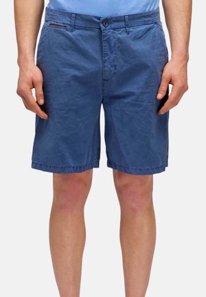 Uomo che indossa pantaloncini blu casual fino al ginocchio e una camicia azzurra, in piedi con le braccia rilassate ai lati.