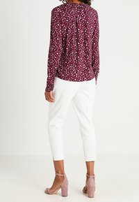 Blusa a maniche lunghe con motivo floreale in bordeaux scuro, abbinata a pantaloni bianchi a sigaretta e sandali con tacco a blocco rosa chiaro.