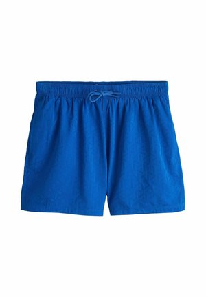 Bărbat tânăr purtând o cămașă neagră cu mâneci scurte și nasturi, maiou alb, pantaloni scurți albaștri și ochelari de soare, stând în picioare lângă un perete bej texturat.