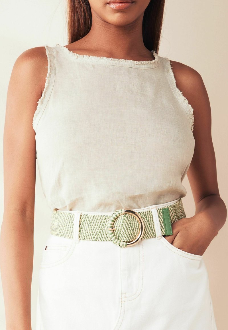 Beige Leinen-Tanktop mit fransigen Kanten, kombiniert mit einem gemusterten grün-weißen Woven-Gürtel mit einer runden Goldschnalle. Weiße Jeans sind sichtbar.