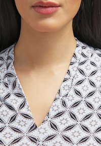 Mönstrad blus med V-ringning, gjord av lätt tyg med ett geometriskt mönster i svart och beige på en ljusblå bakgrund.