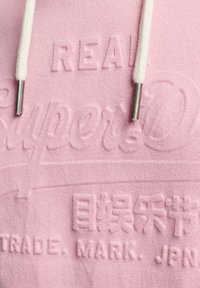 Sudadera rosa con un logo texturizado impreso, cordones blancos en contraste y aglets metálicos. Superficie de tela suave y lisa.