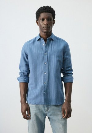 Hombre con camisa azul de botones con rayas blancas finas y jeans azul claro, de pie contra un fondo blanco liso.