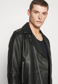 Chaqueta de biker de cuero negro con un gran cuello, cremallera diagonal y bolsillos laterales. Textura suave con detalles metálicos en plata.