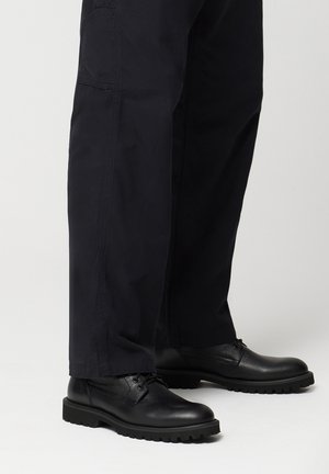 Czarne skórzane buty na sznurowadła z teksturowaną cholewką, gumową podeszwą i solidnym wzorem bieżnika, połączone z ciemnymi spodniami.