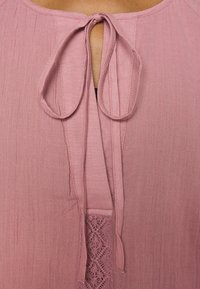 Gros plan d’un vêtement rose poudré avec un nœud en tissu noué et un détail de panneau en dentelle à l’encolure.