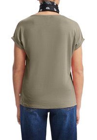 Osoba w oliwkowej koszulce z podwiniętymi rękawami, niebieskich dżinsach i czarnej chustce z białymi wzorami na szyi, odwrócona plecami.