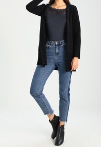 Musta pitkä neuletakki tummansinisen pitsiyläosan päällä, yhdistetty korkeavyötäröisiin sinisiin farkkuihin ja mustiin nilkkureihin; rento istuvuus.