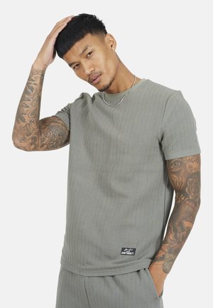 Mężczyzna z tatuażami w szarym, dopasowanym, teksturowanym krótkim rękawie i spodniach, dotykający głowy jedną ręką, na białym tle.