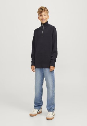 Pull à demi-zip marine en tissu texturé, associé à un jean bleu clair et des baskets décontractées. Un jeune se tient les mains relaxées.