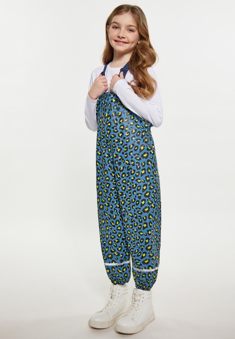 Salopette con stampa leopardata blu e accenti verdi; polsini elasticizzati, spalline regolabili e vestibilità ampia. Indossata sopra una camicia a maniche lunghe bianca.