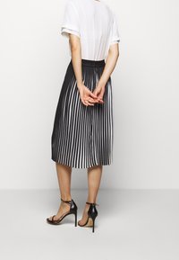 Femme debout les mains jointes derrière le dos, portant un haut blanc à manches courtes, une jupe plissée à rayures verticales noir et blanc, et des talons hauts noirs.